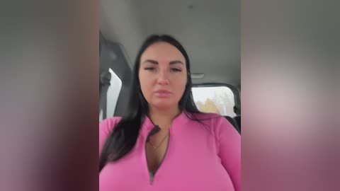 A video of a middle-aged woman with long black hair and fair skin, wearing a pink, long-sleeved top, seated in a car, with blurred car windows in the background.