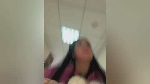 A blurred video of a woman with long hair in a pink and black striped shirt lying on a bed, with a white blanket and a window in the background.
