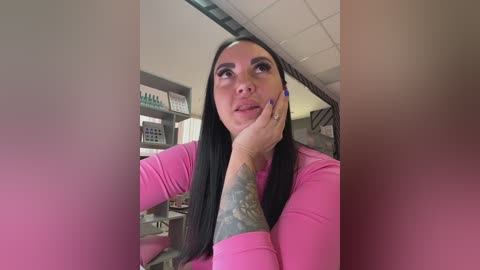 Video of a dark-haired woman with a tattooed arm in a bright pink shirt, looking thoughtful in a medical office with shelves and white ceiling tiles.