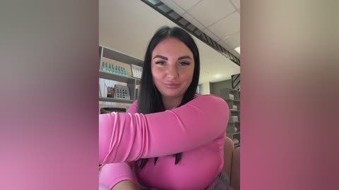 A video of a smiling woman with long black hair and light skin, wearing a tight pink top, hugging a large pink cushion. The background features shelves with books and a staircase.
