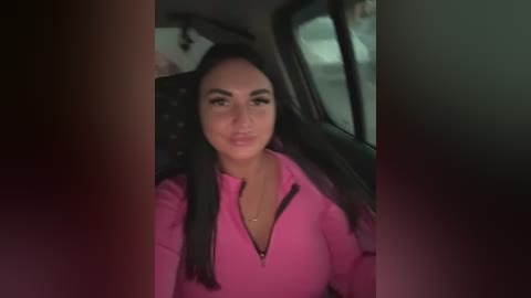 Video of a young woman with long dark hair, wearing a bright pink zip-up hoodie, seated in a dimly lit car.