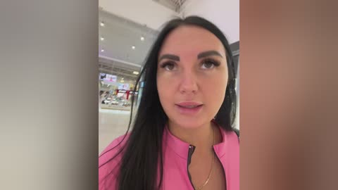 Video of a young woman with long black hair, wearing a pink jacket, speaking into a microphone in a modern indoor setting with blurred background.