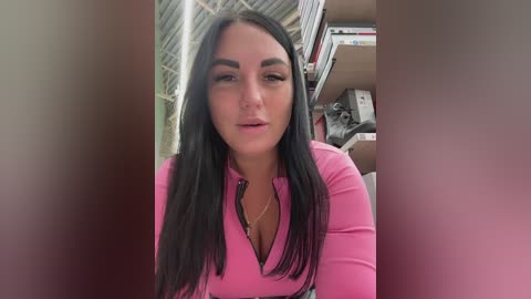Video of a young woman with long black hair, wearing a pink long-sleeve top, indoors near shelves and boxes, looking slightly seductive.