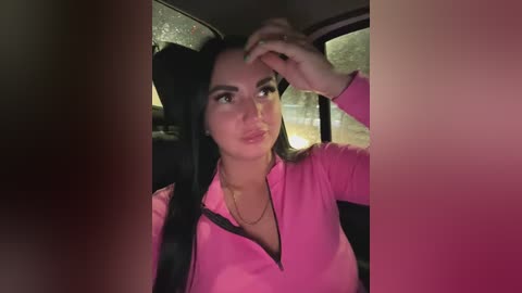 Video of a woman with long black hair, wearing a bright pink zip-up top, sitting in a car at night, with raindrops on the window.