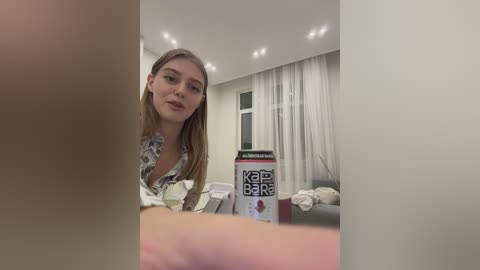 Media: Video of a young woman with light skin, straight blonde hair, wearing a floral blouse, sitting on a sofa in a modern living room with white curtains and recessed lighting.