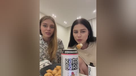 Media: Video of two young women, one with long blonde hair and the other with long black hair, eating chicken nuggets from a \"KABOBRA\" box in a bright, modern kitchen.