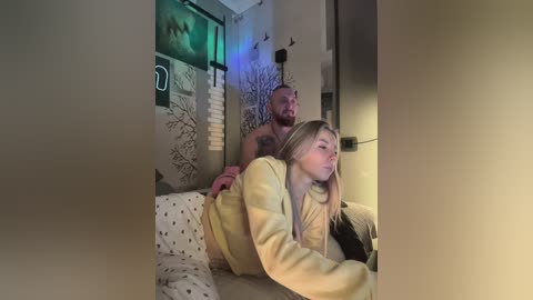 Video of a nude muscular man with short hair, standing behind a blonde woman in a yellow sweater, on a bed with patterned sheets, in a dimly lit room.