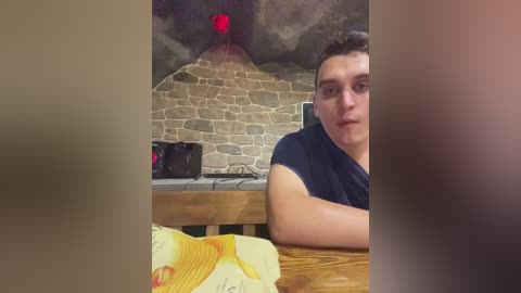 Media: Video of a young man with short dark hair, wearing a black T-shirt, seated at a wooden table with a yellow napkin and fork. The background features a stone wall and a red lava lamp, creating a cozy, rustic atmosphere.