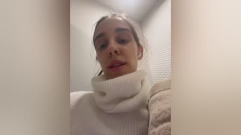 Media: A video of a young woman with light skin, dark hair, and brown eyes, wearing a white turtleneck sweater, taken from a low angle, creating a distorted perspective.