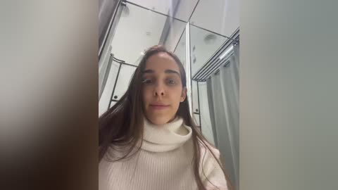 Media: A video of a young woman with long brown hair and light skin, wearing a white turtleneck sweater, standing in a modern, well-lit bathroom with mirrors and glass shower doors.