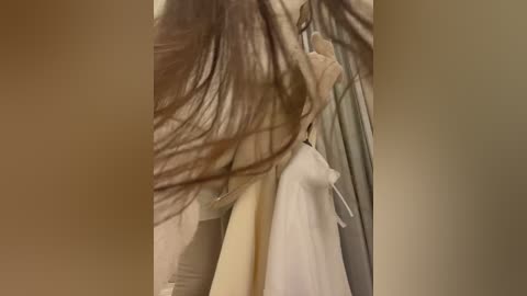 Media: Video of a woman with long, dark brown hair, wearing a beige trench coat, bending over. The background is blurred with a brownish tone, suggesting an indoor setting.