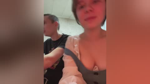 Video of a young Asian woman with short black hair and a medium build, wearing a low-cut, white top with a gray vest, sitting next to a Caucasian man in a black T-shirt, in a dimly lit room.