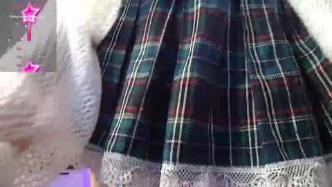 Media: A close-up video of a woman's plaid skirt with green, red, and white stripes, and a white lace-trimmed edge. The background is blurred, focusing on the skirt's intricate detailing.