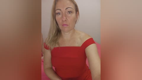 Video of a Caucasian woman with blonde hair, wearing a red off-shoulder dress, sitting against a pink background. Her facial expression is neutral, and the image is slightly blurred.