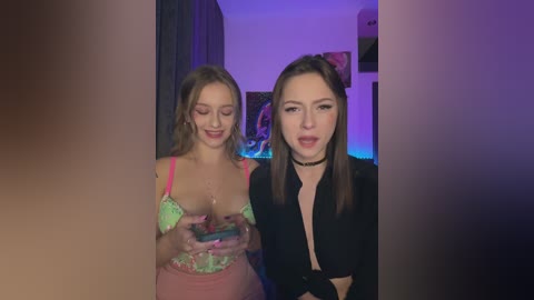 Media: Video of two young women, one with light skin, green bra, and pink skirt, holding a phone, the other with darker skin, black choker, and black top, in a dimly lit room with purple lighting and framed artwork in the background.