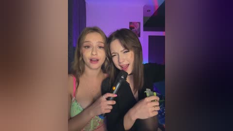 Media: Video of two young women, one with light skin and green bra, the other with fair skin and black top, singing into a microphone in a dimly lit room.