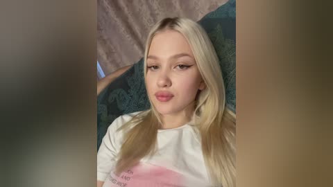 Media: Video of a young, fair-skinned woman with long blonde hair, wearing a white T-shirt, lying on a teal-patterned bedspread. The background features a blurred, light-colored wall and a pink curtain.