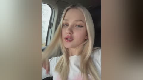 Media: Video of a young, light-skinned, platinum blonde woman with long hair, wearing a white t-shirt, sitting in a car. Her mouth is slightly open, and she appears to be in a relaxed state.