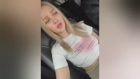 Media: A video shows a young blonde woman with fair skin and closed eyes, wearing a white crop top and beige pants, reclining in a car seat.