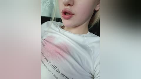 Media: Video of a young, fair-skinned woman with blonde hair, wearing a white T-shirt with a pink stain on the chest, lips slightly parted.