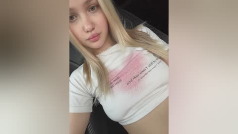 Media: Video of a young Caucasian woman with long blonde hair, fair skin, and small breasts, wearing a tight white T-shirt with a pink stain. She sits in a car with a dark interior.