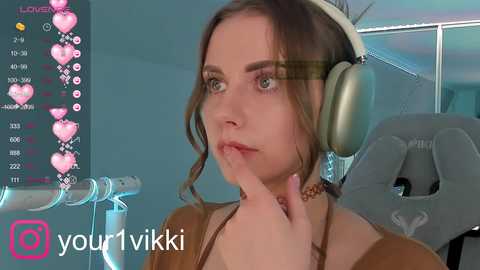 Media: A video of a young woman with light skin and brown hair, wearing headphones and a brown top, in a streaming setup with a gray gaming chair.
