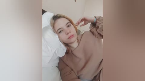 A video of a young woman with light skin and blonde hair, lying on a bed, wearing a beige sweater. She has a contemplative expression. The background is blurred, focusing on her face.