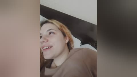 Video of a young woman with fair skin and shoulder-length red hair, smiling, wearing a brown shirt, in a bedroom with white walls and a black headboard.
