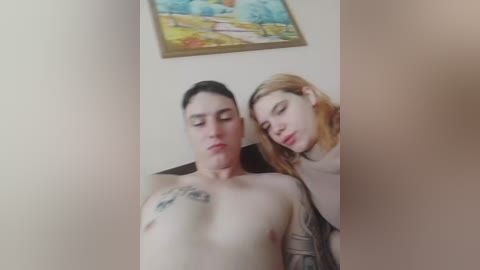Video of a topless, tattooed man with fair skin lying on a bed, with a fair-skinned, blonde woman resting her head on his chest. Background features a framed landscape painting.