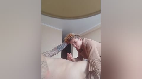Video of a fair-skinned, curly-haired woman in a beige dress, kneeling on a beige couch, holding a large, erect penis with intricate tattoos.