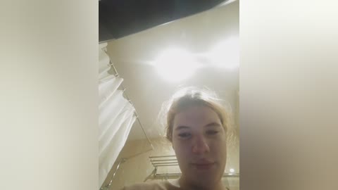 Video of a young, fair-skinned woman with light brown hair, looking downward, captured in a dimly lit bathroom with a white shower curtain and beige walls.