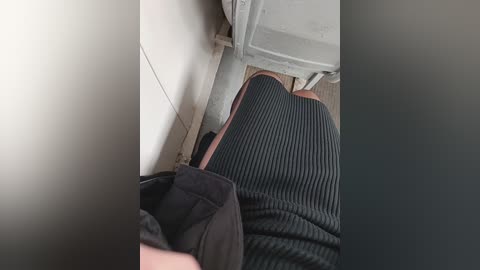 Video of a person's legs in black ribbed socks and brown shoes, sitting on a gray bench in a tiled bathroom with white walls and a visible drain.