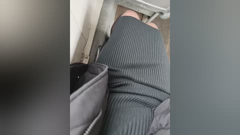 A video shows a person wearing a gray, ribbed knit dress and black leggings, sitting on a toilet in a public restroom. The background features a tiled wall and wooden flooring.