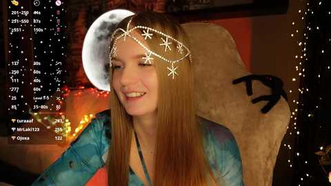 A video of a smiling young woman with long, straight, light brown hair, wearing a turquoise top and a silver moon headband, seated in a dimly lit, cozy room with a large stuffed animal.