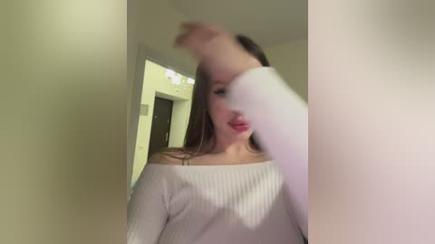 Media: Video of a woman in a white off-shoulder sweater, with her face partially obscured by a hand, in a dimly lit hallway with pale green walls and a closed door.