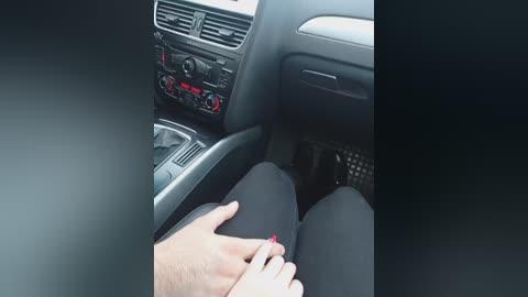 Video of a person with light skin, wearing black pants, sitting in a car, holding another person's hand, showing red nail polish. The car interior includes a black dashboard with a steering wheel and various controls.