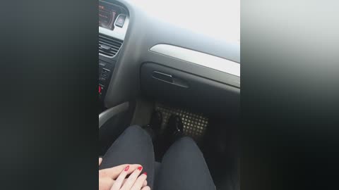 Video of a car interior with a person's legs and feet visible, wearing black pants and red-painted toenails. The steering wheel and dashboard are visible, with a beige seat cover and dark upholstery.