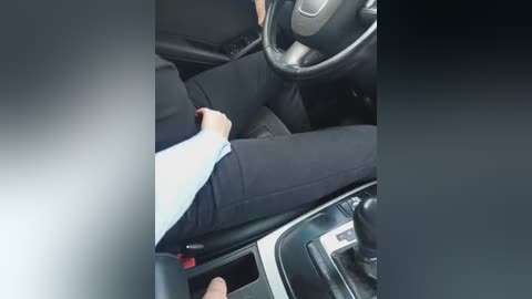 A video shows a person's leg and hand inside a car, wearing dark pants and a light blue shirt, with a black steering wheel in the background.