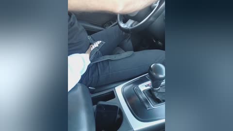 Video of a person wearing black pants, sitting in a car, with the car's gearshift and steering wheel visible, showing a casual, everyday scene.