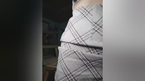 Video of a person's lower body, partially covered by a gray plaid blanket, in a dimly lit room with a bookshelf and cluttered background.