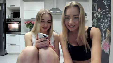 Media: Video of two smiling, blonde women in black tank tops, sitting in a modern kitchen with white cabinets, black stove, and chalkboard wall.