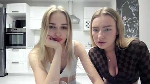 Media: Video of two blonde Caucasian women with fair skin in a modern kitchen. One wears a white tank top, the other a black checkered shirt, both looking thoughtful. White cabinets, stainless steel appliances, and a chalkboard with a sketch in the background.