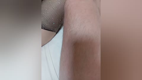 A close-up video of a person's upper arm with a mesh sleeve and white tank top, showing fair skin and subtle shadows, set against a blurred beige background.