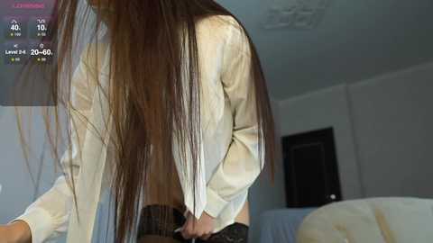 Media: Video of a woman with long brown hair, wearing a white blouse and black shorts, bending over in a modern, minimally decorated bedroom.