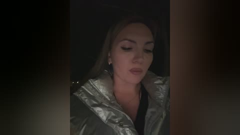 Video of a woman with light skin, wearing a shiny silver raincoat, standing in a dimly lit room with a dark background, focusing on her face.
