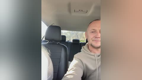 Media: Video of a bald, light-skinned man with a slight smile, wearing a gray hoodie, sitting in the driver's seat of a car with black leather seats.