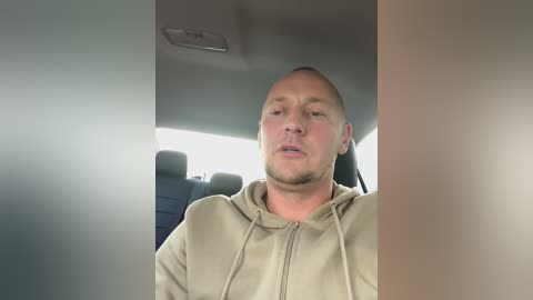 Media: Video of a bald, middle-aged Caucasian man in a beige hoodie, sitting in the backseat of a car, looking slightly annoyed.