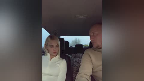 Media: Video of a blonde woman in a white sweater and a bald man in a beige sweater seated in a car, window view of a cloudy sky.