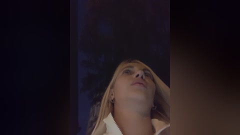 Media: Video of a blonde woman with fair skin, wearing a white sweater, looking upward with a serene expression. Background is dimly lit, possibly a dark room.