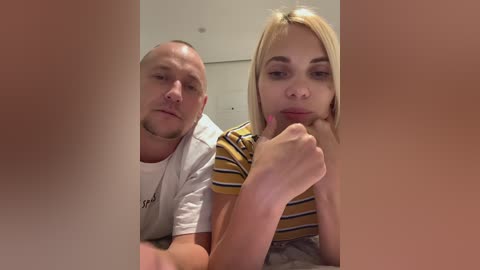 Media: Video of a Caucasian man and woman with fair skin, both wearing casual clothes. The man is bald, wearing a white shirt, and the woman has blonde hair and a yellow and white striped shirt.